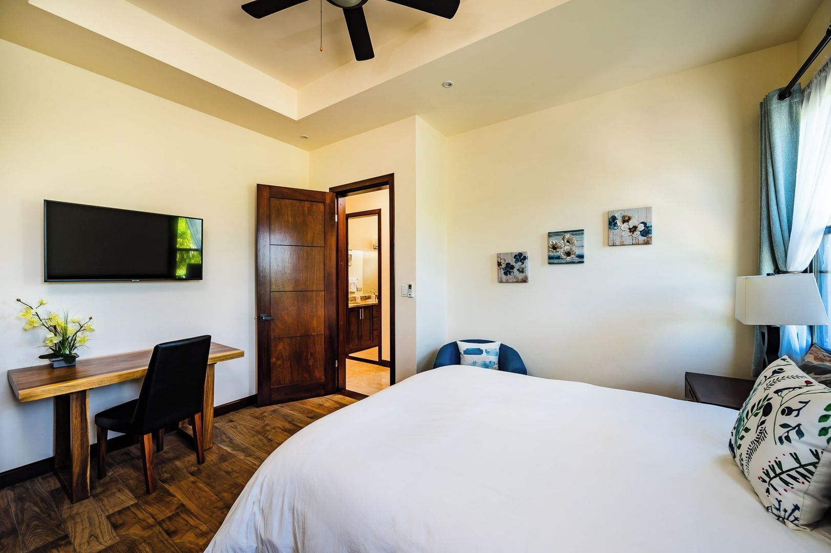 Quarto de hotel com cama, escrivaninha, cadeira, televisão e porta para o banheiro.