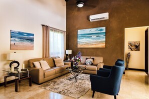 Ein Wohnzimmer mit einem beigefarbenen Sofa, einem Glastisch, einem blauen Sessel und einem großen Gemälde, das eine Strandansicht zeigt.