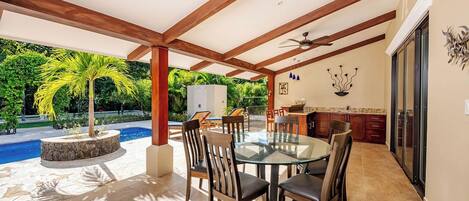 A covered outdoor dining area with a glass table and chairs, a ceiling fan, and a view of a swimming pool and greenery.