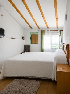 A bedroom with a large bed, wooden beams on the ceiling, a television on the wall, and a window with curtains. - 🌿 Iñaki's Refugio – Nature, tranquility, and vacations with your dog (Mijangos)