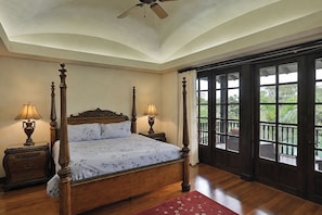 A four-poster bed with a floral bedspread, two bedside tables with lamps, a red rug, and a balcony with a view of greenery. - 3-Bedroom Villa in Hacienda Pinilla (Pinilla)