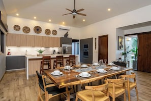 A modern kitchen with wooden cabinets, a central island, and a dining area with a large wooden table and chairs. - Casa Pochote | Hacienda Pinilla | Golf and Beach (Pinilla)