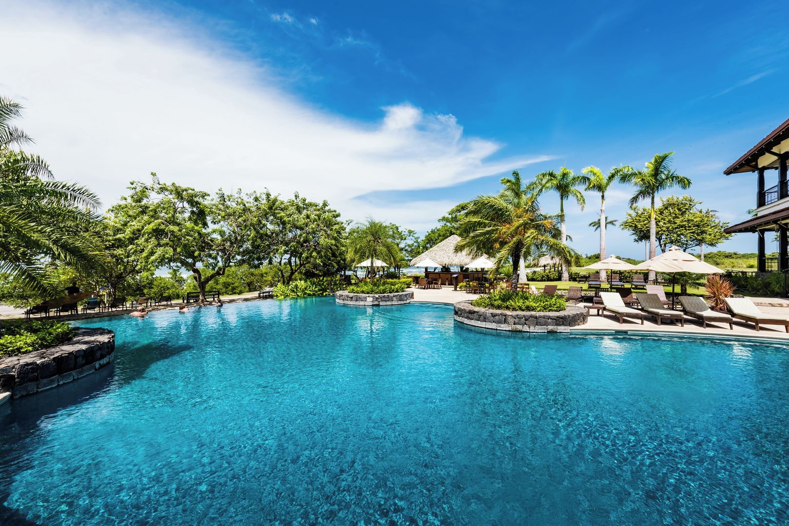 Une piscine d’un bleu éclatant, entourée de chaises longues et de parasols, avec des palmiers et une structure à toit de chaume en arrière-plan.