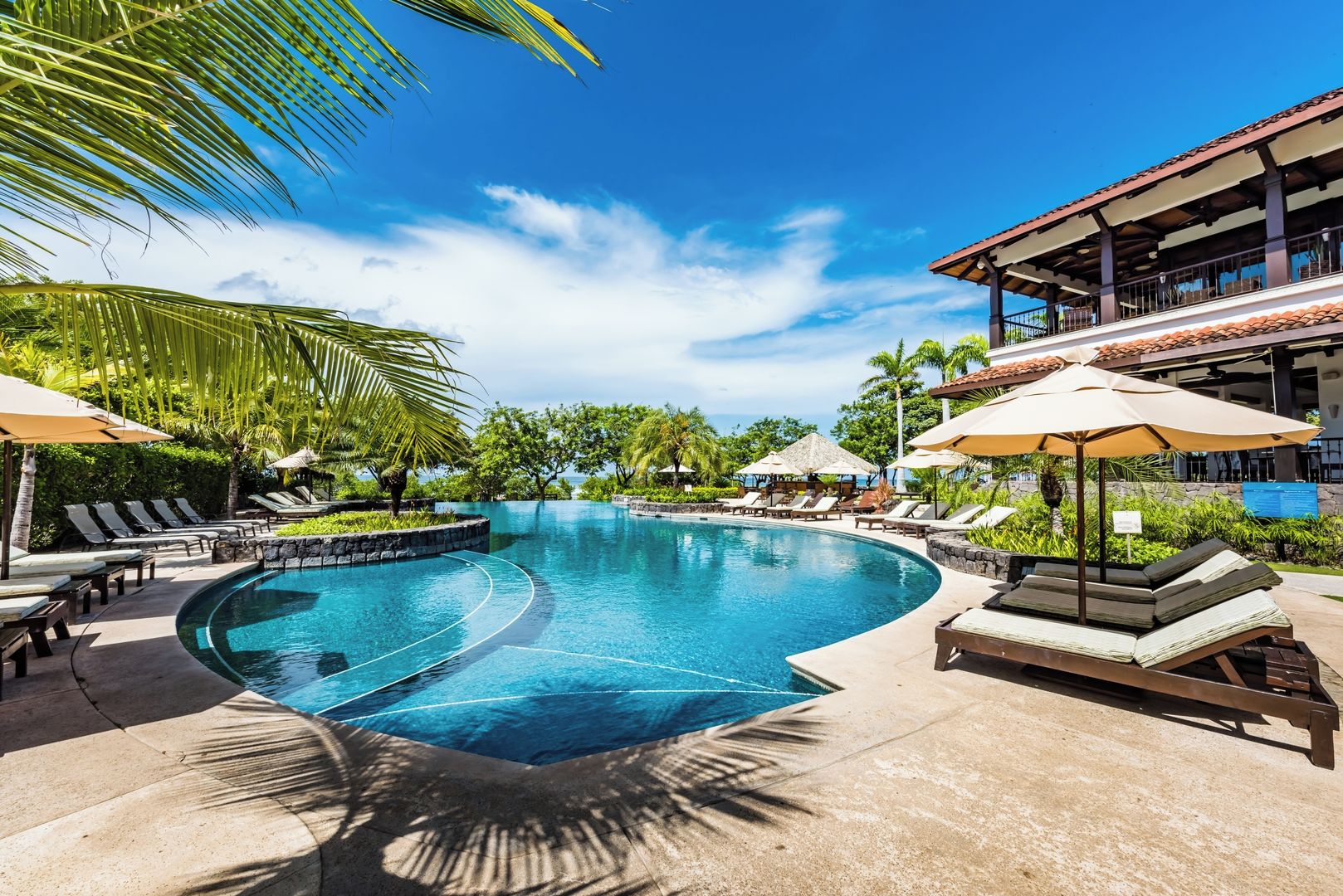 A swimming pool with lounge chairs and umbrellas, surrounded by palm trees and a building with a tiled roof.