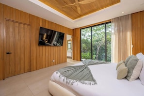 A modern bedroom with a large bed, wooden panel walls, a flat-screen TV, and a balcony with a view of greenery. - Casa Solara | Hacienda Pinilla (Hacienda Pinilla)