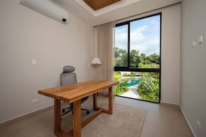 A room with a wooden table, a chair, a large window with a view of a swimming pool and greenery, and an air conditioning unit. - Casa Solara | Hacienda Pinilla (Hacienda Pinilla)