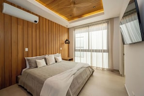 A bedroom with a wooden paneled wall, a bed with a beige bedspread, a ceiling fan, and a TV mounted on the wall. - Casa Solara | Hacienda Pinilla (Hacienda Pinilla)