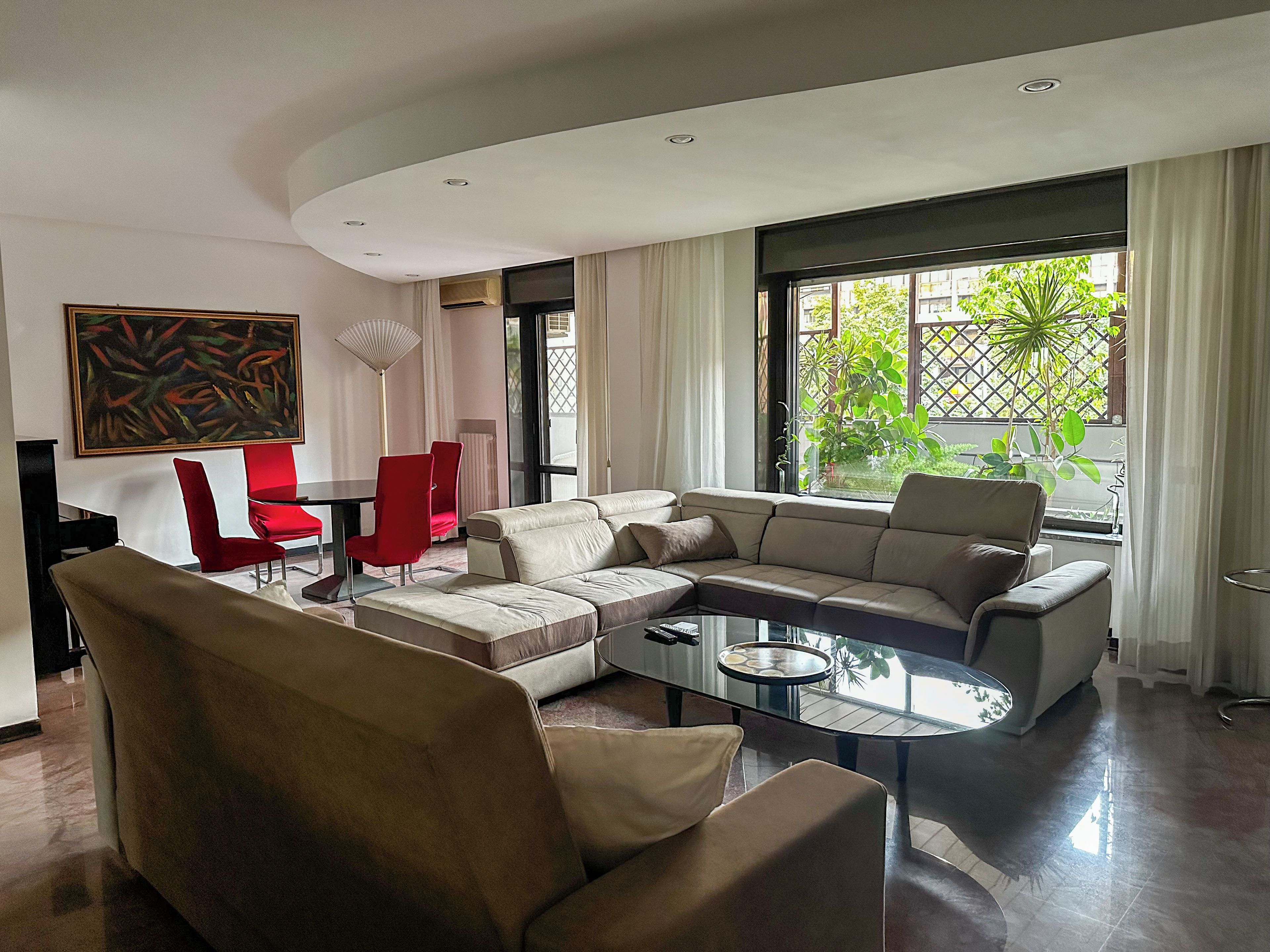 Un salon moderne avec un canapé d’angle, des fauteuils rouges, une table basse en verre et une grande fenêtre donnant sur un paysage verdoyant.
