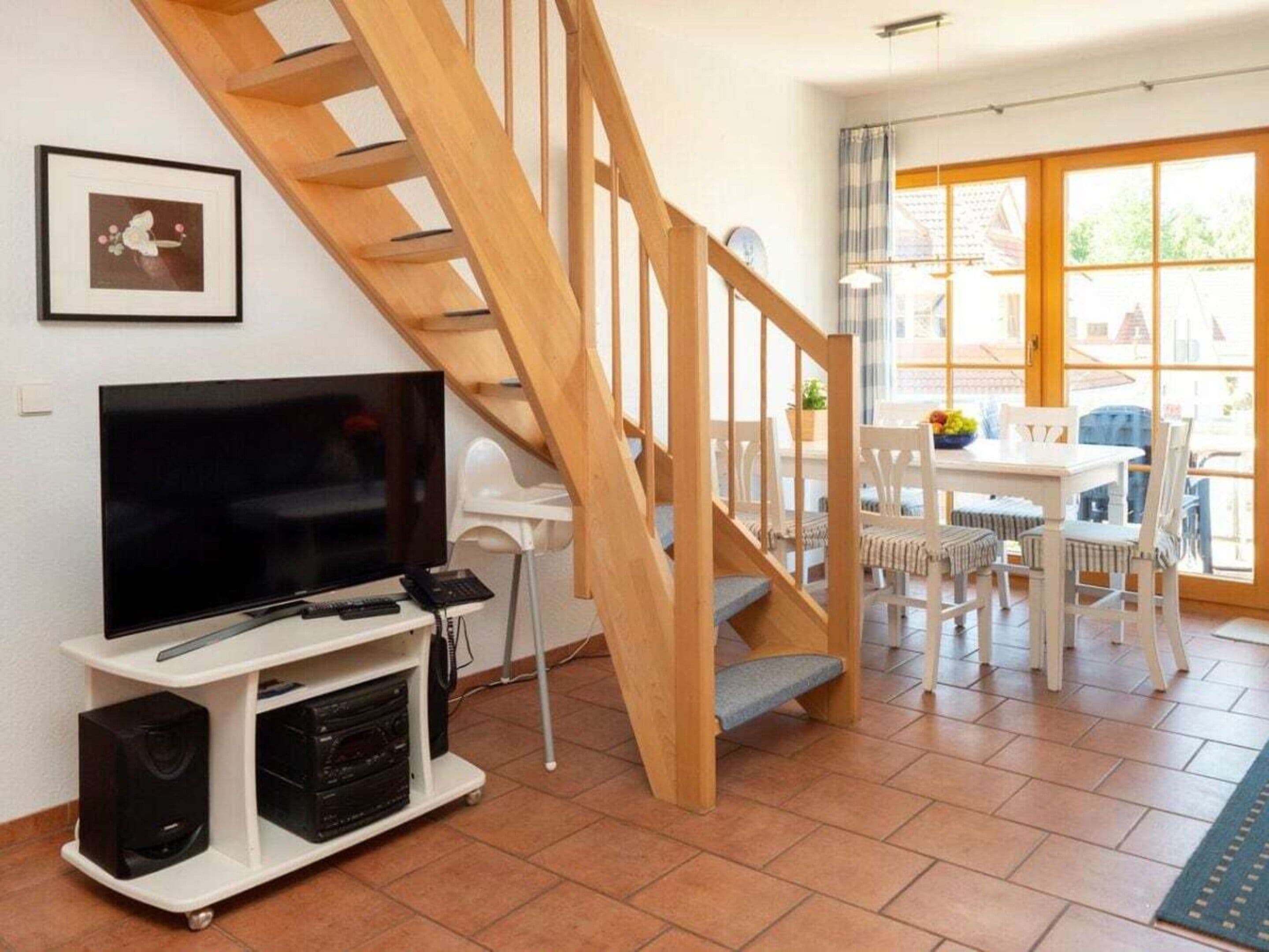 Una sala de estar con una escalera de madera, un televisor, un mueble blanco para entretenimiento y un comedor con mesa y sillas.