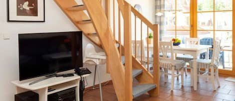 Una sala de estar con una escalera de madera, un televisor, un mueble blanco para entretenimiento y un comedor con mesa y sillas.