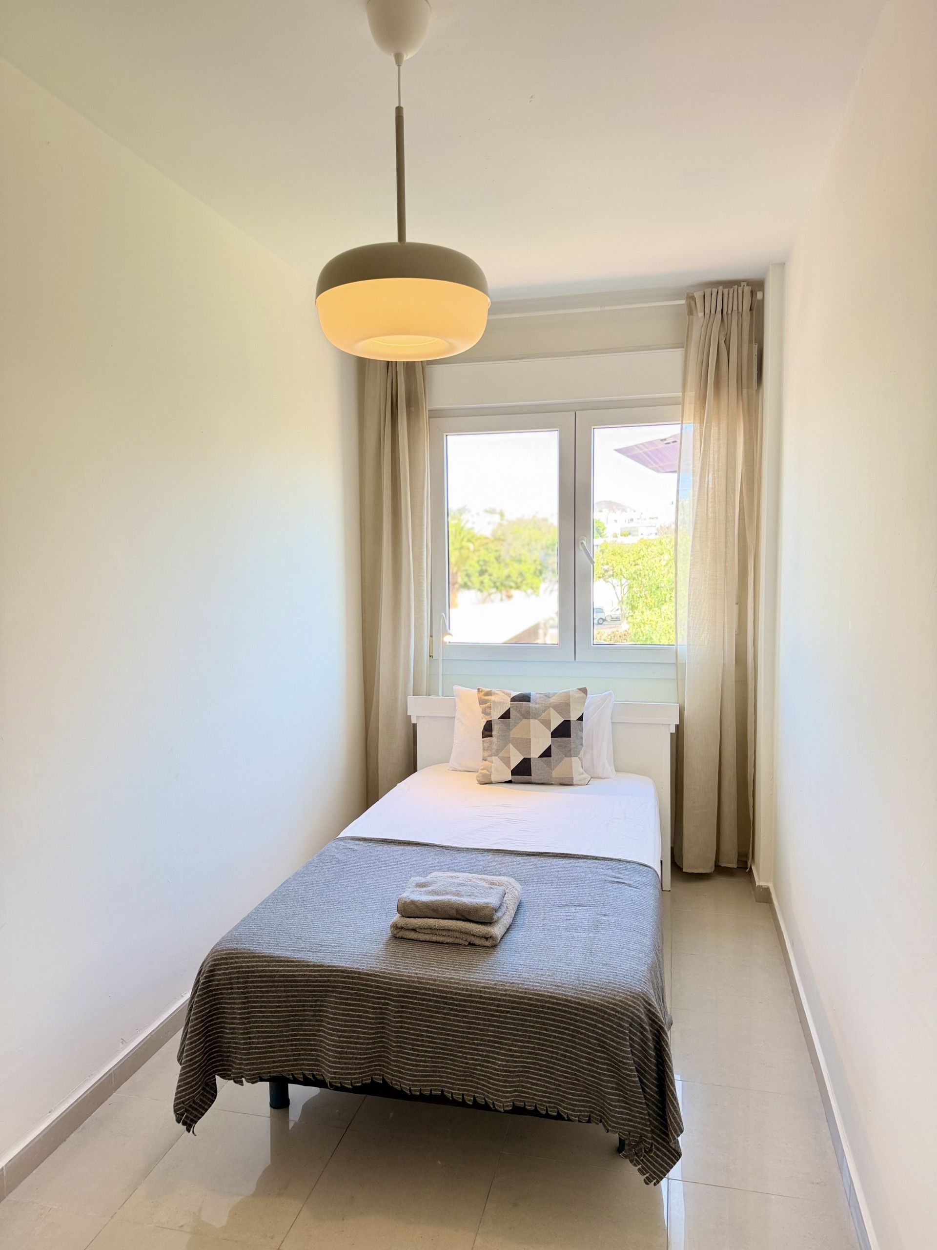 A single bed with a striped bedspread, a pillow, and a folded towel on the floor. A window with curtains is visible, and a hanging light fixture is above the bed.
