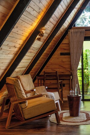 Habitación en ático con vigas de madera, una silla de madera, una mesa con una botella y una ventana con cortinas.