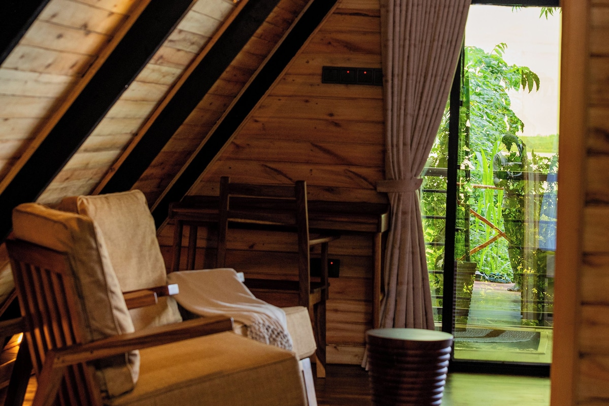 A cozy room with a sloped ceiling, a wooden chair, a small table, and a view of greenery outside.