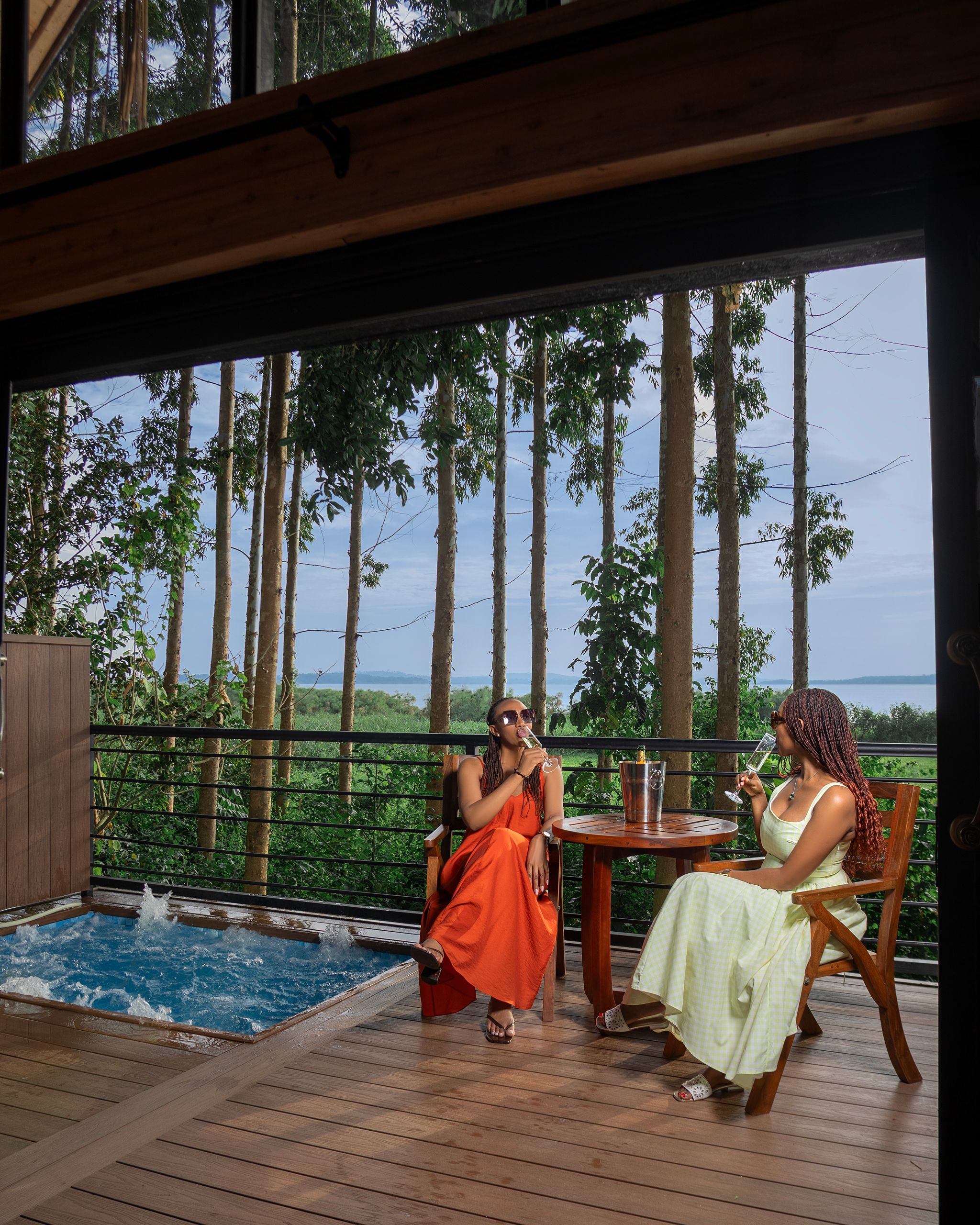 A wooden deck with a pool, two people sitting at a table, and a view of trees and water.