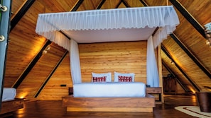 Una cama con dosel de madera con cuatro columnas, ropa de cama blanca y almohadas estampadas en una habitación con paredes y techos inclinados de madera.