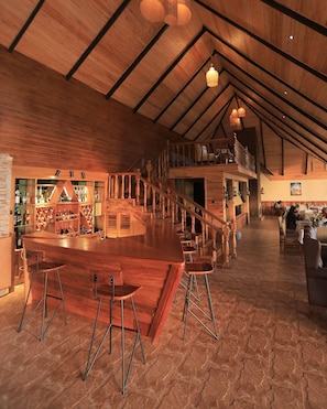 Un amplio espacio tipo bar con techo alto y vigas de madera, que incluye una disposición de taburetes y una estantería para vinos.