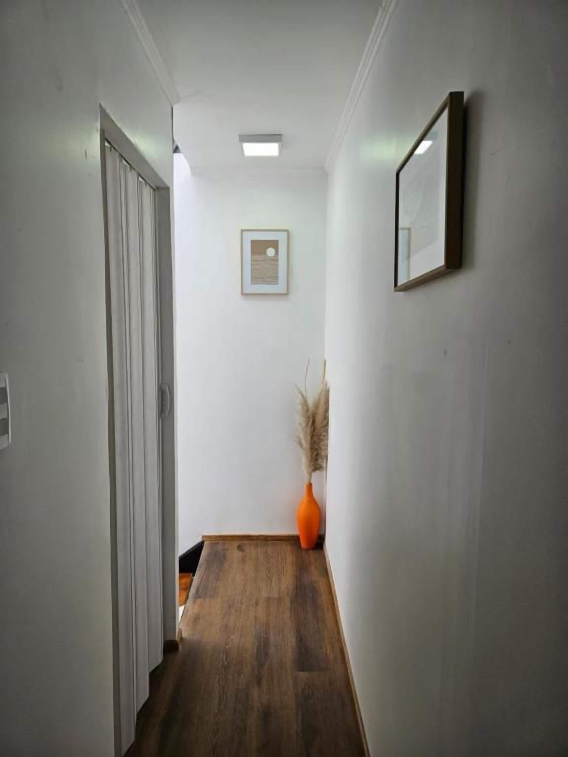 Un pasillo con piso de madera, una puerta a la izquierda, un cuadro en la pared y un jarrón con plantas secas.