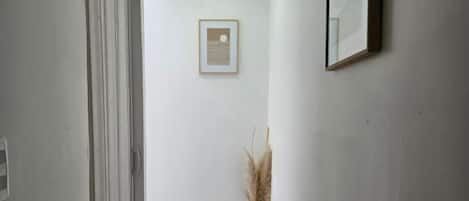 A hallway with a wooden floor, a door on the left, a framed picture on the wall, and a vase with dried plants.