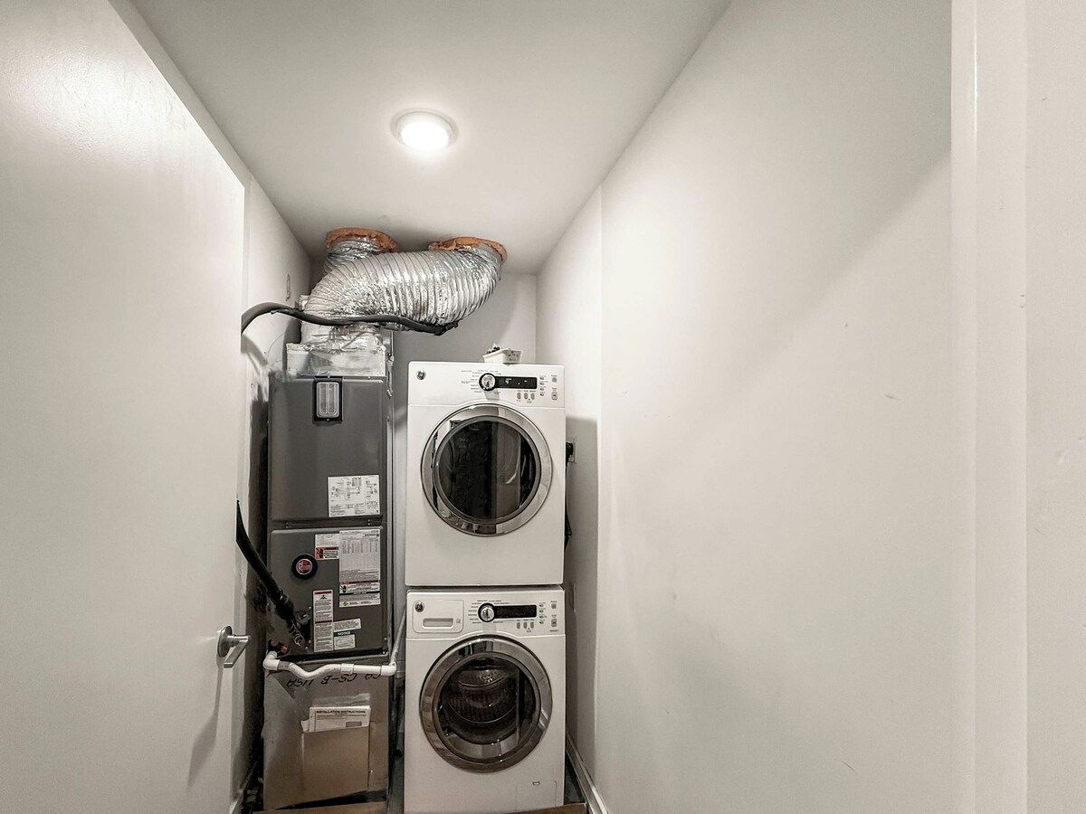 A laundry room with two stacked washing machines and a dryer, a vent above the machines, and a door on the left.
