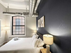 A hotel room with a large bed, a window with a view of a brick building, a dark wall with artwork, and a lamp on the nightstand. - Luxury 2BD | 4 Beds | Northern Liberties (Philadelphia)
