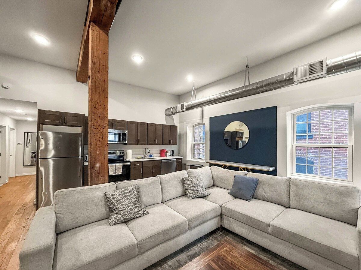 A modern living room with a sectional sofa, a kitchen in the background, and a large window with a view.