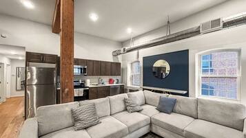 A modern living room with a sectional sofa, a kitchen in the background, and a large window with a view.