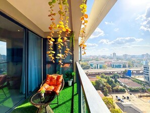 Un balcón con vistas a la ciudad, una silla con cojines anaranjados y hojas artificiales colgantes.
