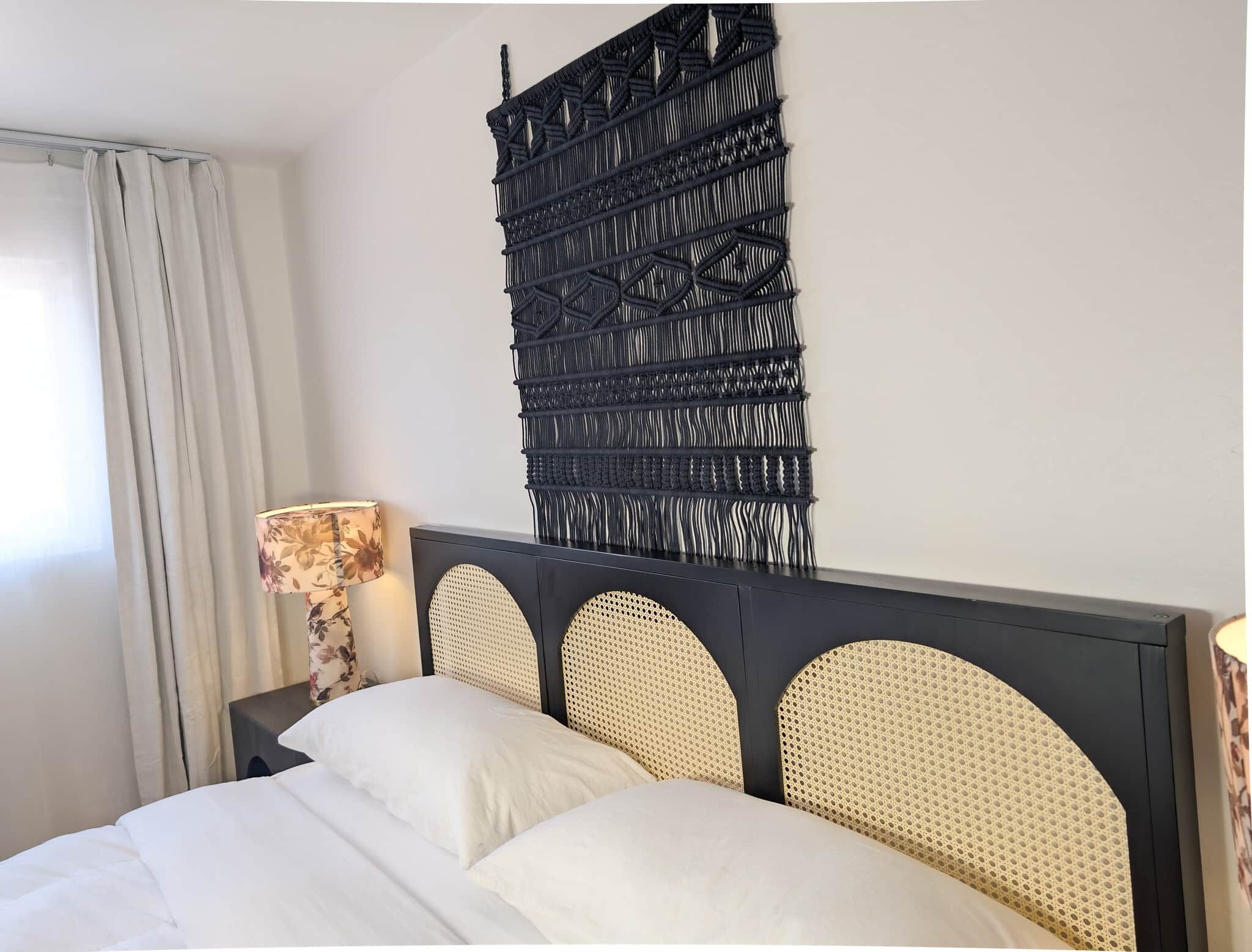 Una cama con ropa de cama blanca, un cabecero oscuro con diseños tejidos y un macramé colgado en la pared.