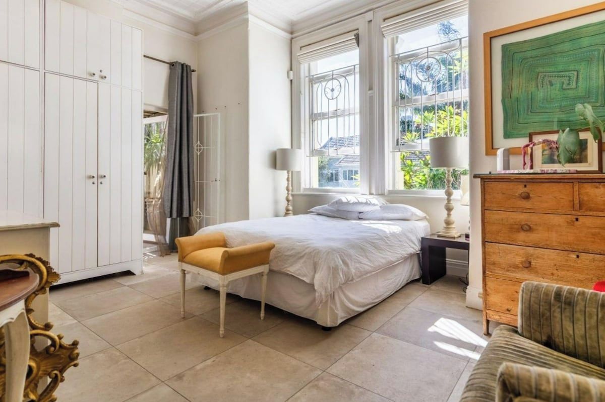 Ein Schlafzimmer mit einem Bett, einer Holzkommode, einem Sessel und einem Fenster mit Blick auf Grünflächen.