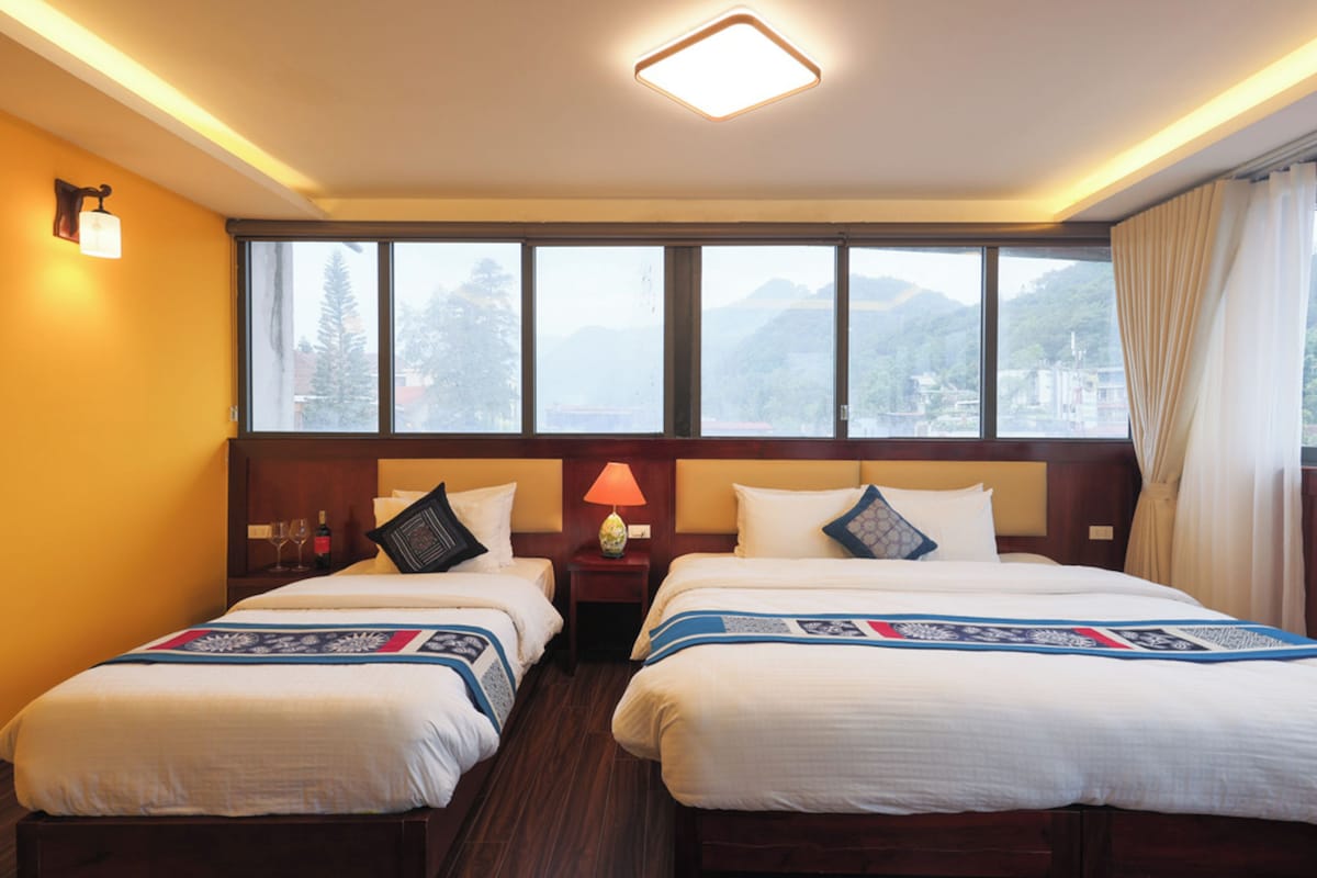 Een hotelkamer met twee bedden, een nachtkastje met een lamp en uitzicht op de bergen door grote ramen.