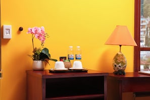 Een kamer met een gele muur, een houten bureau, een potorkidee, twee waterflessen, een lamp en een kleine plant.