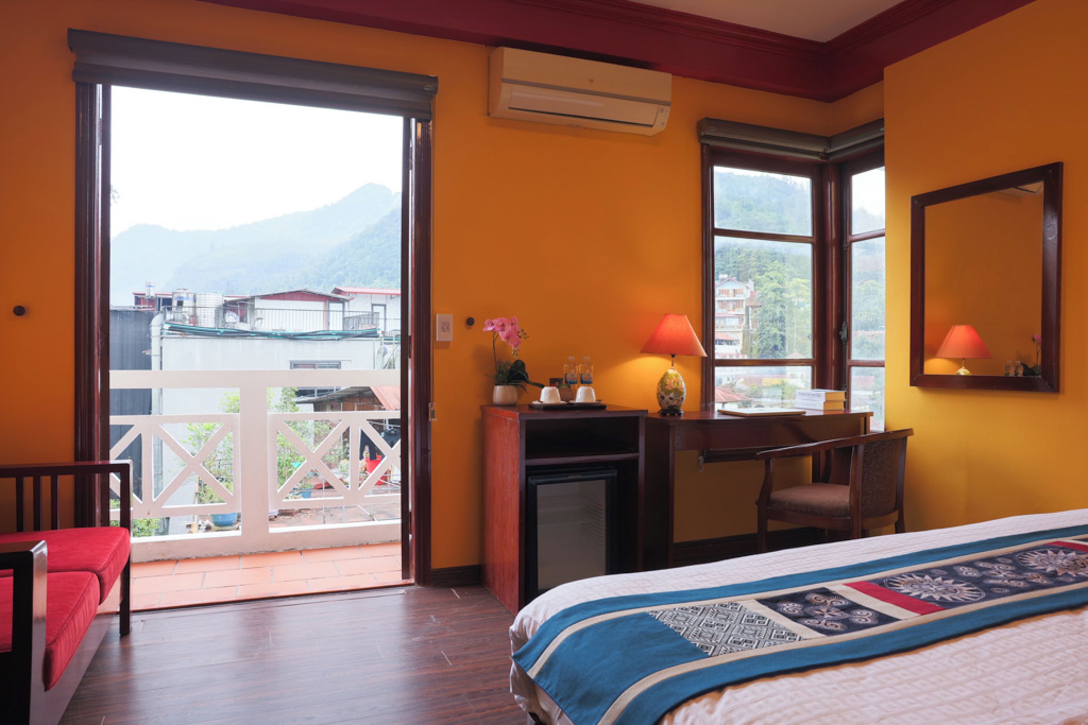 A hotel room with a bed, desk, chair, and a balcony with a view of mountains.