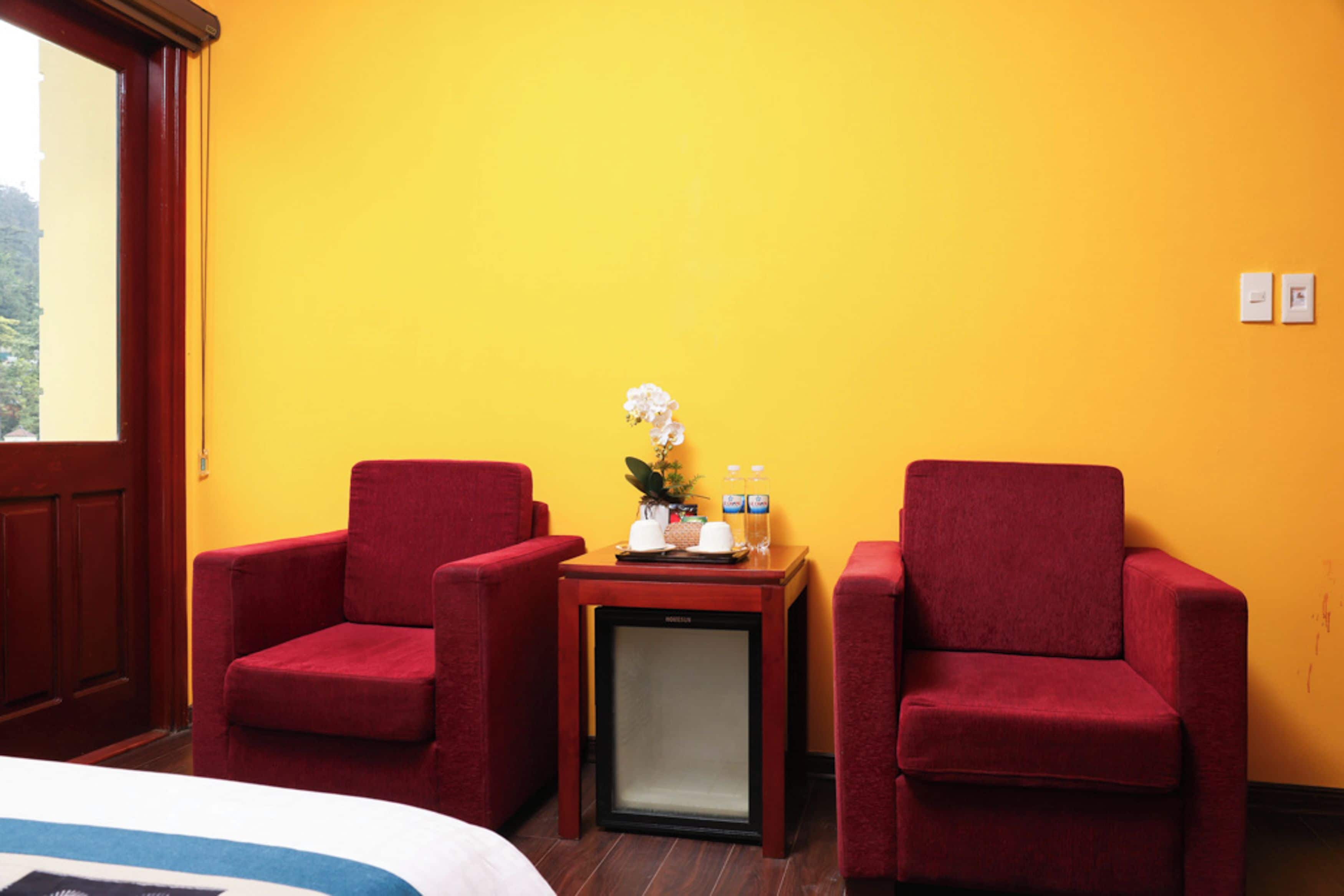 A hotel room with two red armchairs, a small table with a plant, a refrigerator, and a bed with a blue and white duvet.