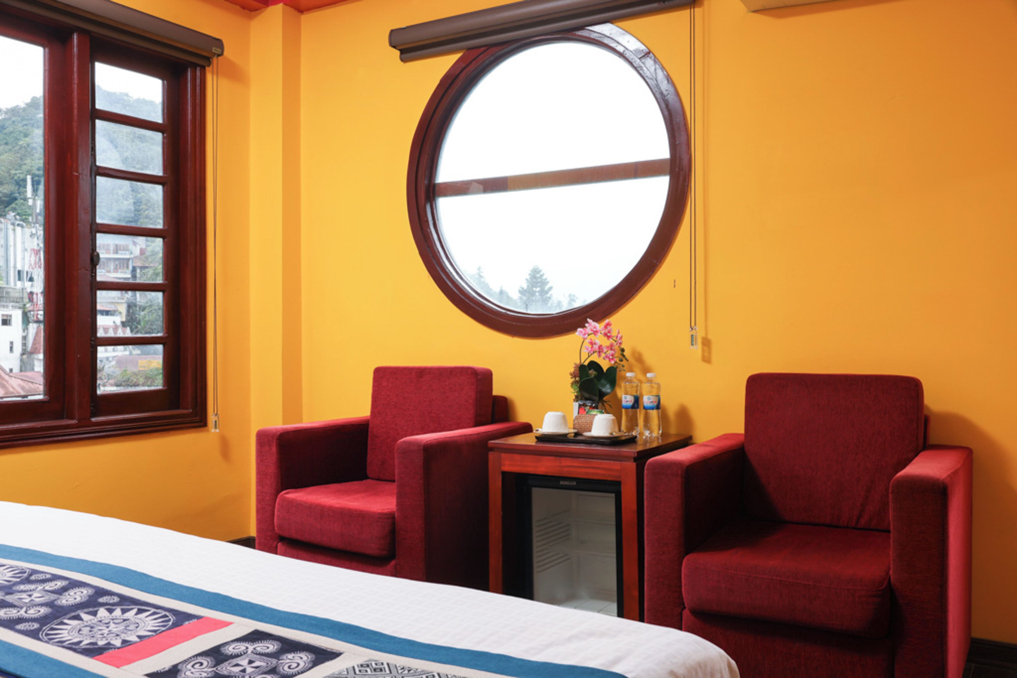 A hotel room with a bed, two red armchairs, a small table with a tray, a round window, and a view of buildings outside.