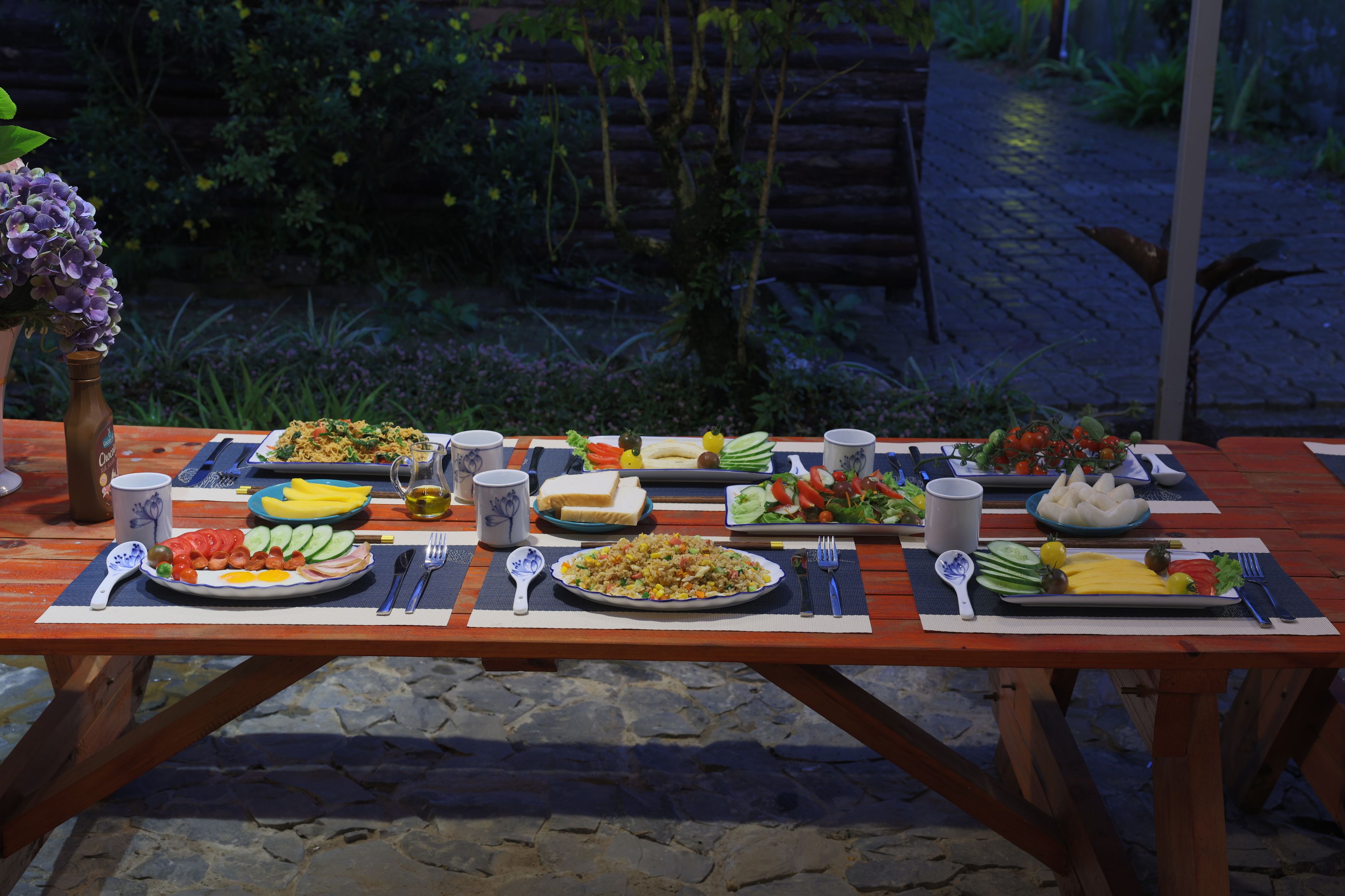 An outdoor dining table set with various dishes, utensils, and a bottle.