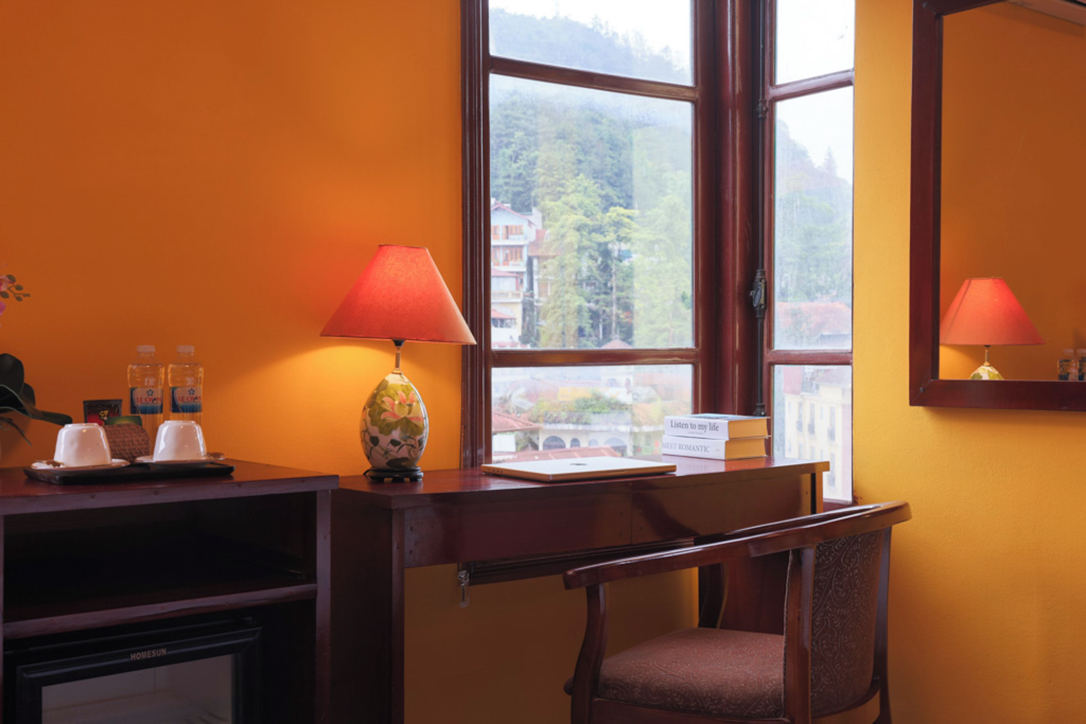 A hotel room with a desk, a chair, a lamp, a small table with cups and a vase, and a window with a view of buildings and trees.