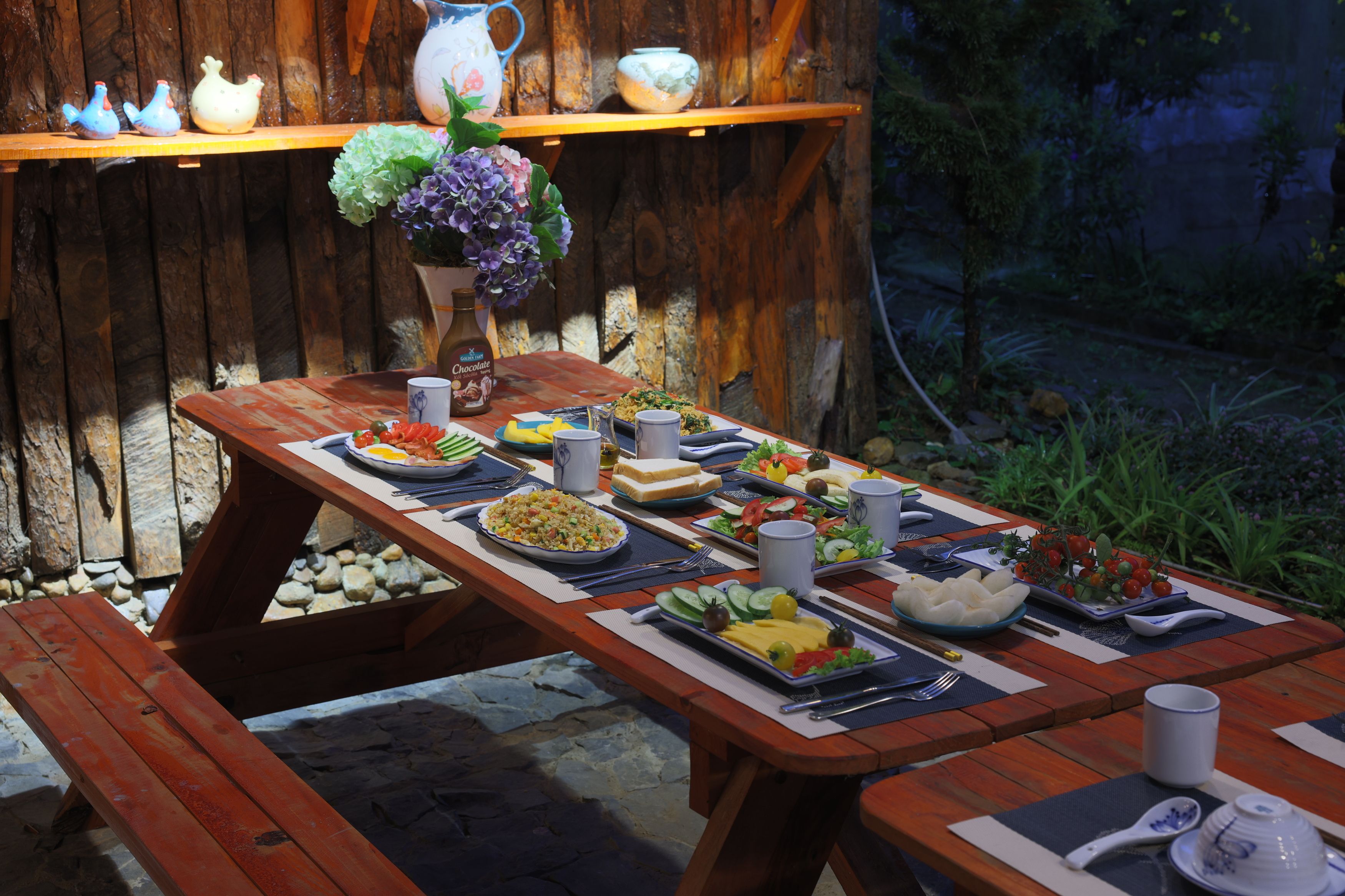 An outdoor dining area with a wooden table set for a meal, surrounded by a rustic wooden fence and a garden.