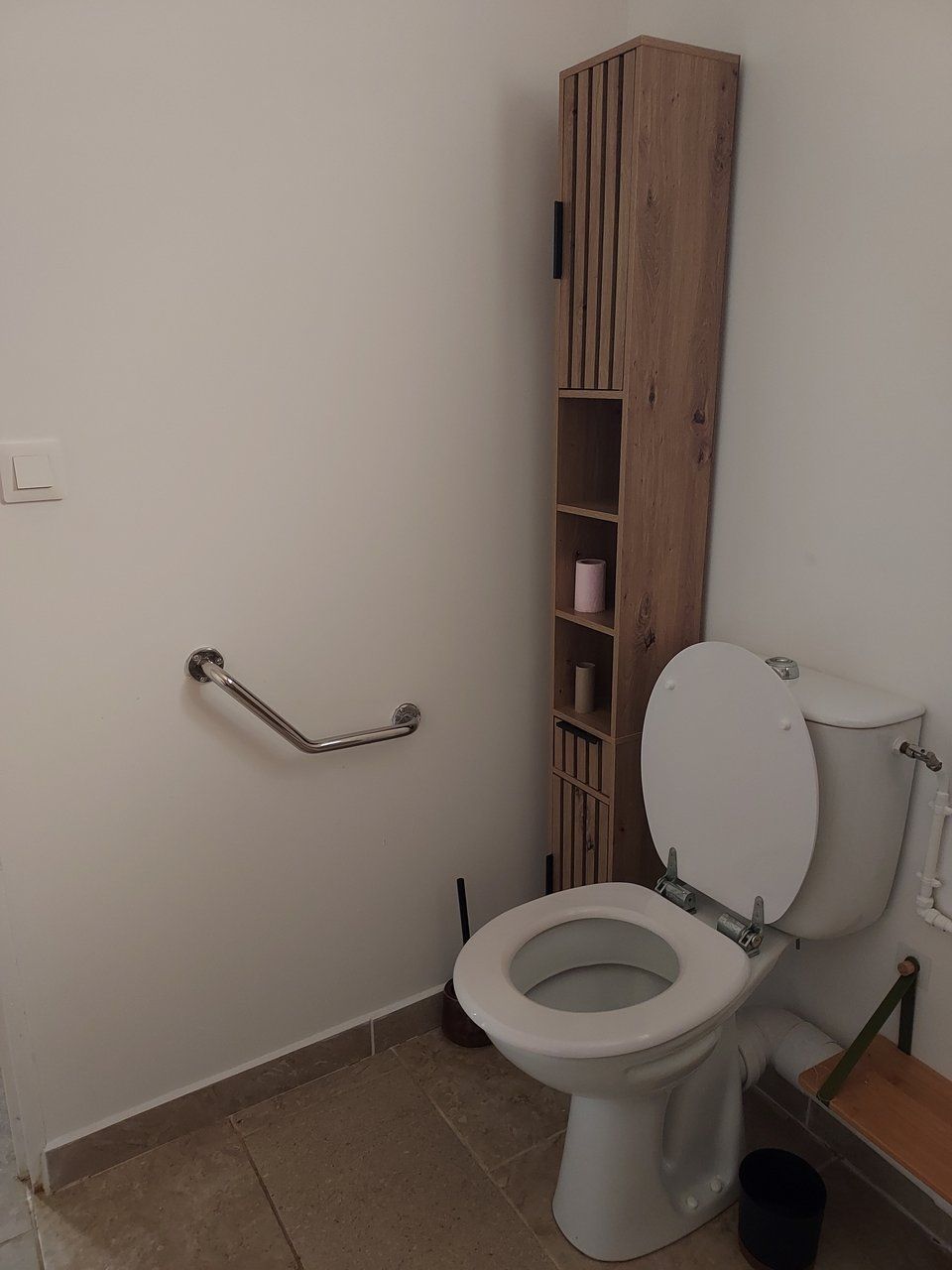 A bathroom with a white toilet, a wooden cabinet with shelves, a grab bar, and a tiled floor.