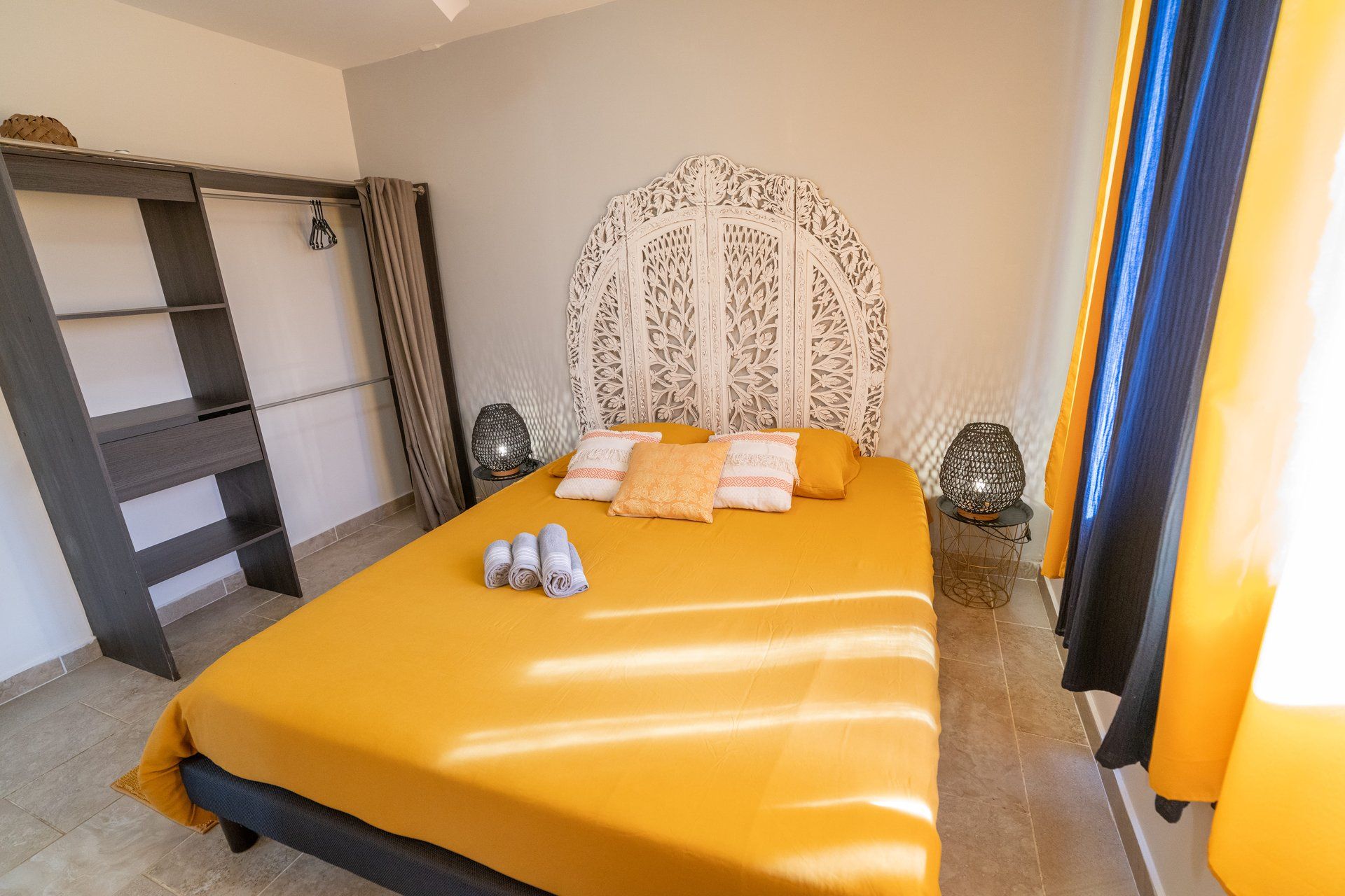 A hotel room with a bed, a headboard with intricate carvings, a small table with a lamp, and a window with curtains.