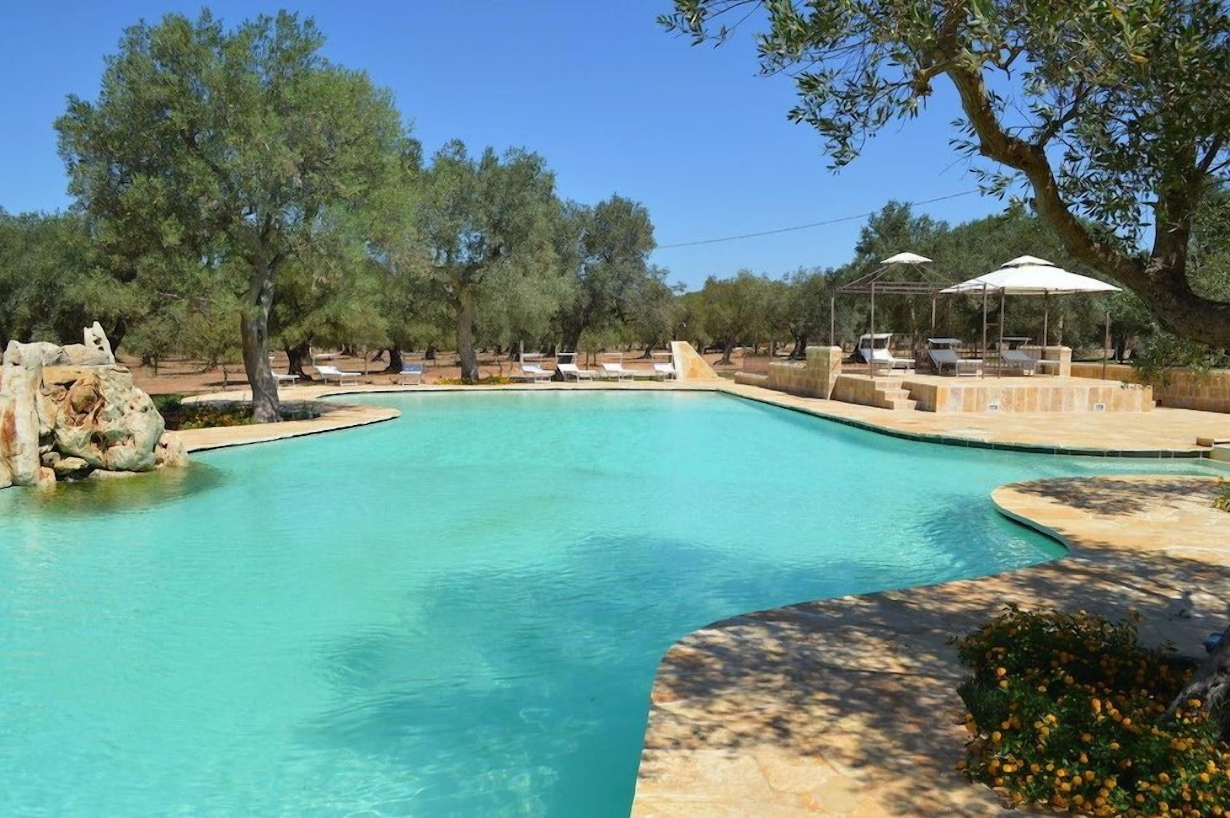 Piscina transparente com cadeiras de praia e guarda-sóis, cercada por árvores e uma estrutura de pedra.