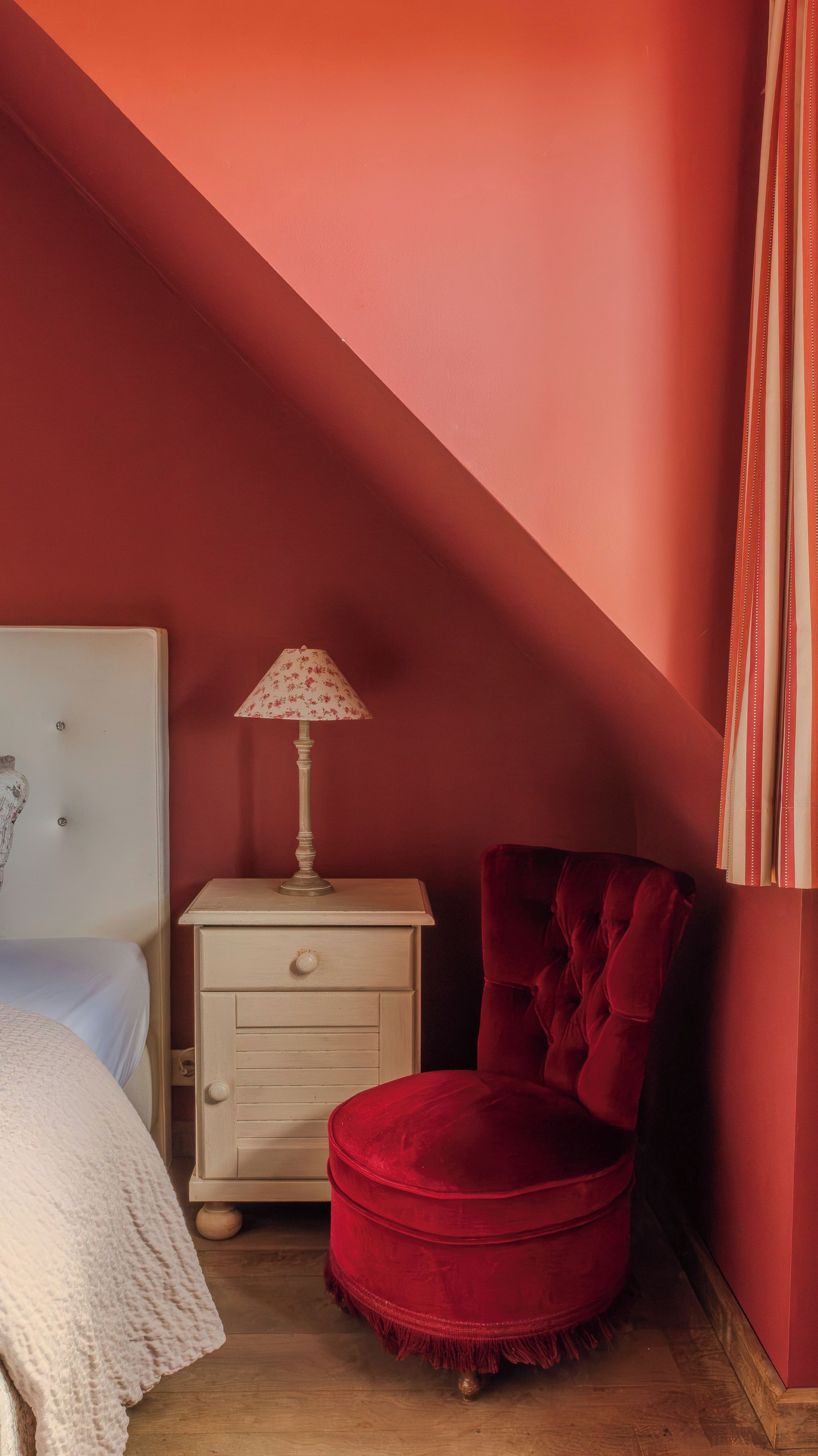 Un dormitorio con una pared roja, una mesita de noche blanca con una lámpara, una silla roja y una cama con una manta blanca.
