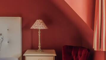 Un dormitorio con una pared roja, una mesita de noche blanca con una lámpara, una silla roja y una cama con una manta blanca.