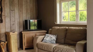 A room with a beige leather sofa, a television on a wooden cabinet, a window with a view of greenery, and a tiled floor.