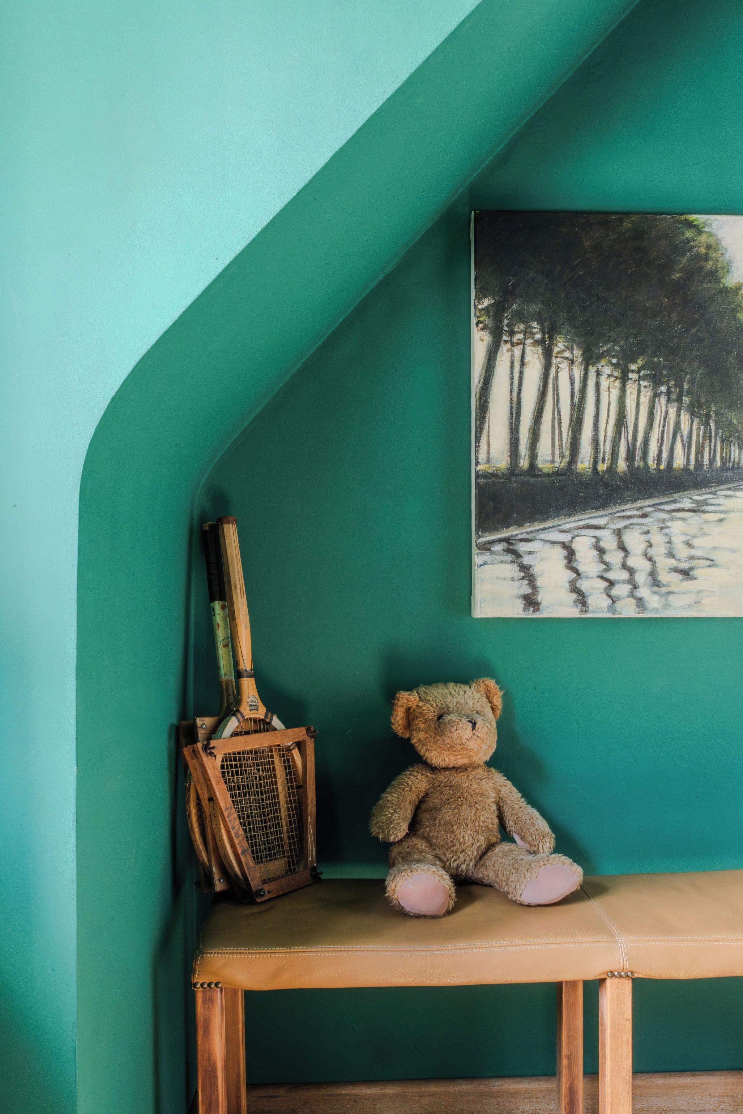 A room with a teal wall, a painting of trees, a wooden bench, and a teddy bear.