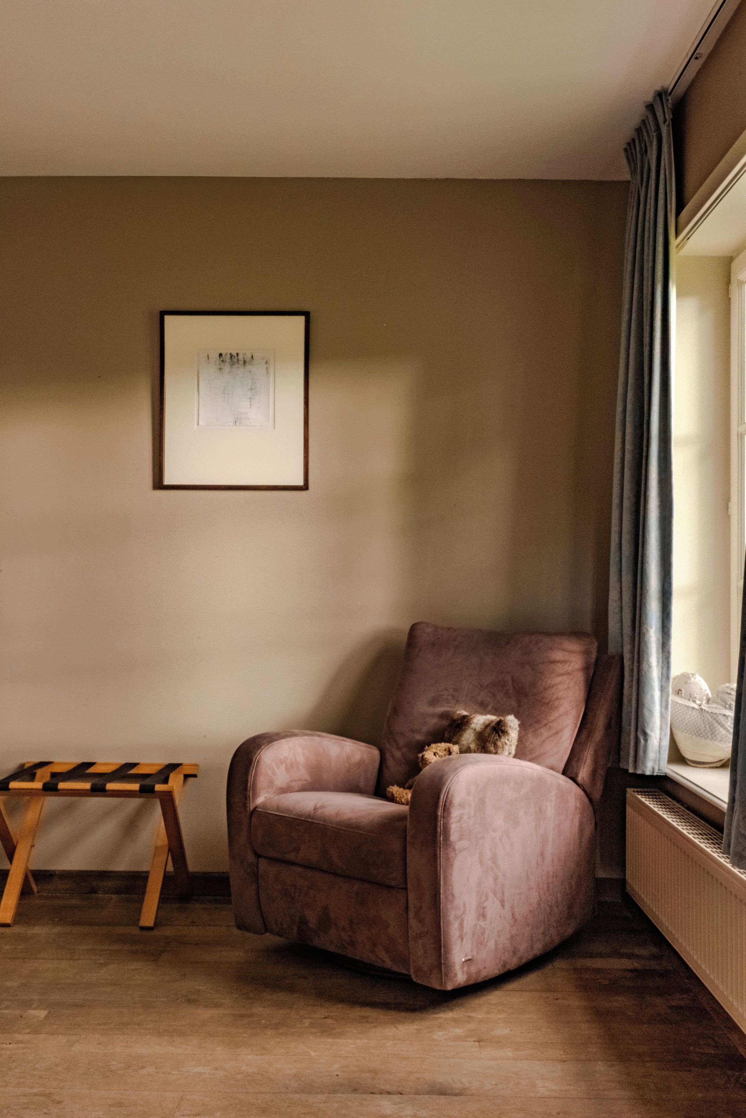 Quarto de canto com poltrona acolchoada, uma pequena mesa de madeira, um quadro na parede e uma janela com cortinas.