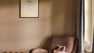 Een hoekappartement met een comfortabele fauteuil, een klein houten tafeltje, een ingelijst schilderij aan de muur en een raam met gordijnen.