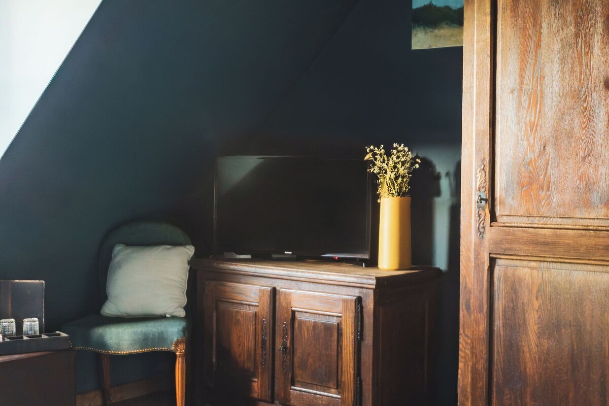 Huone, jossa on kalteva katto, puinen kaappi, tuoli, televisio ja maljakko, jossa on kukkia.
