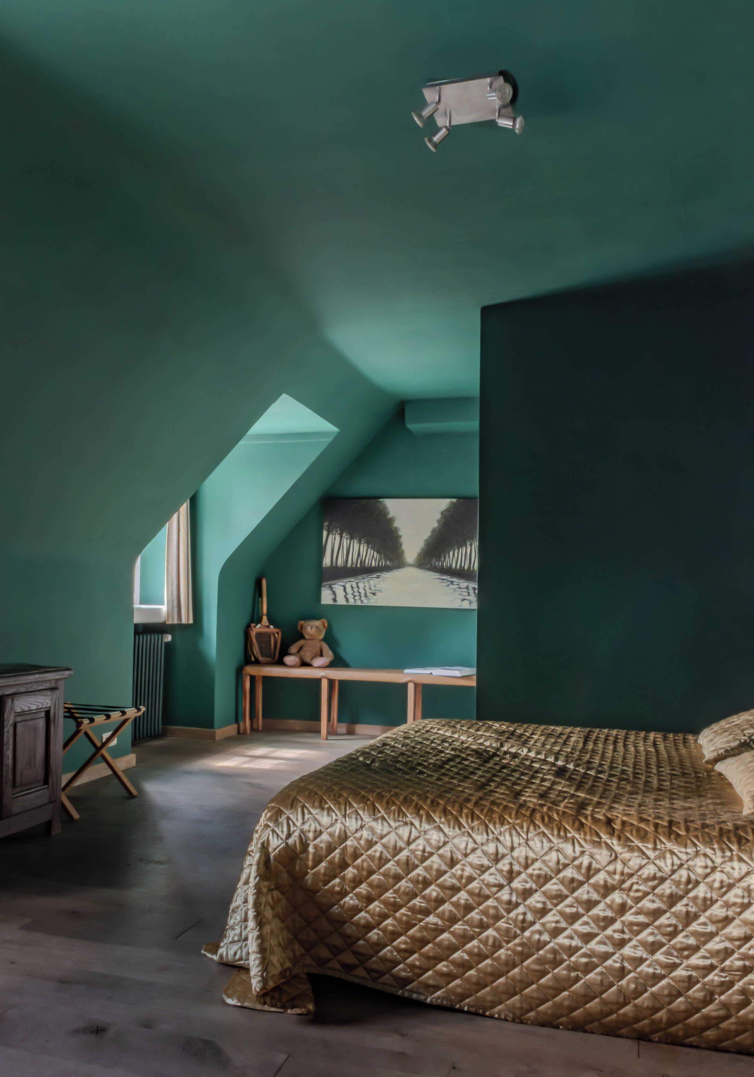 Una camera da letto con soffitto spiovente, un letto con copriletto trapuntato, una panca in legno, un orsacchiotto, una chitarra e un quadro raffigurante alberi.