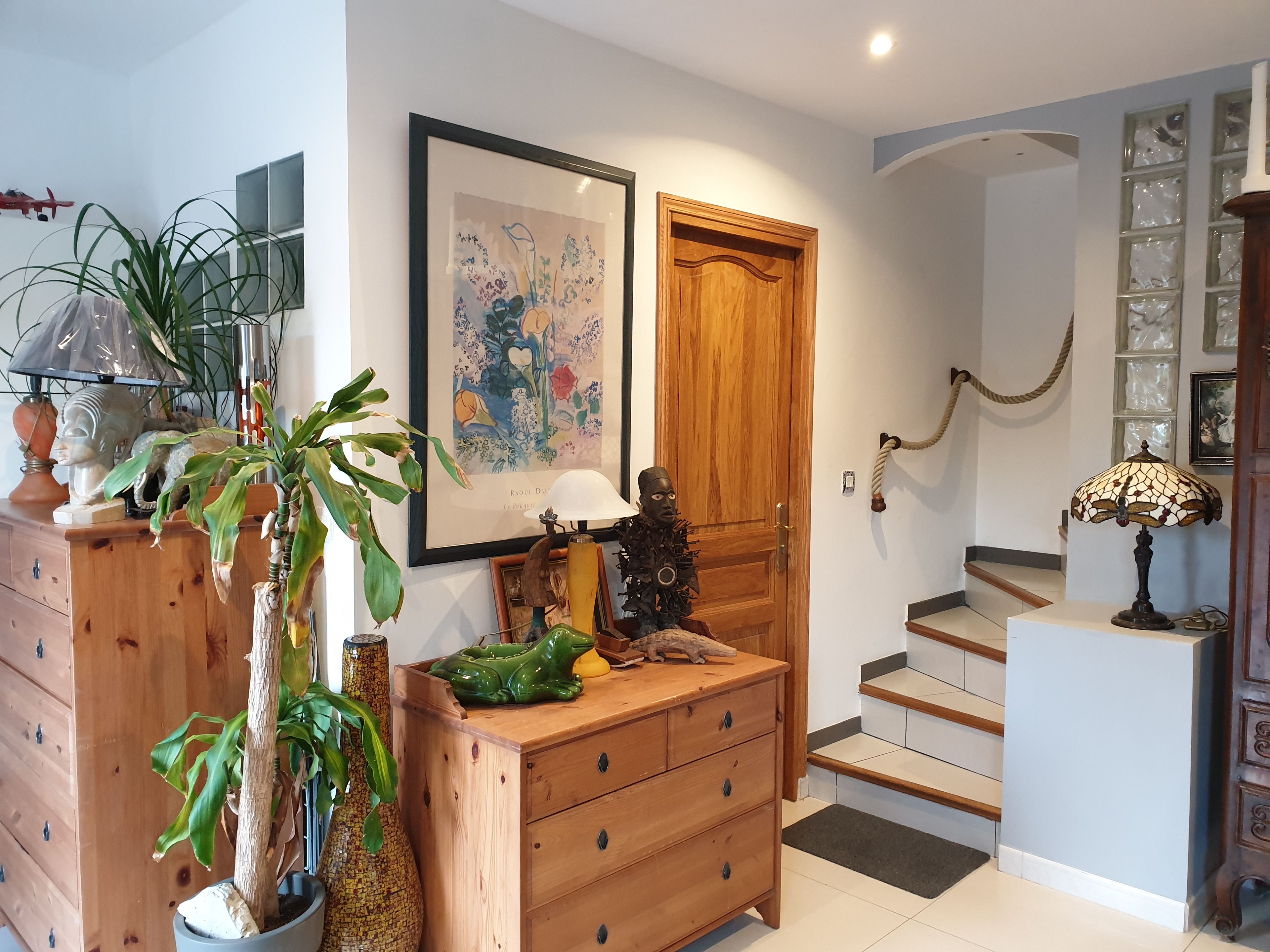 Een goed verlichte gang met een houten trap, een schilderij van bloemen in een lijst en een groene kikkerbeeldje op een houten dressoir.