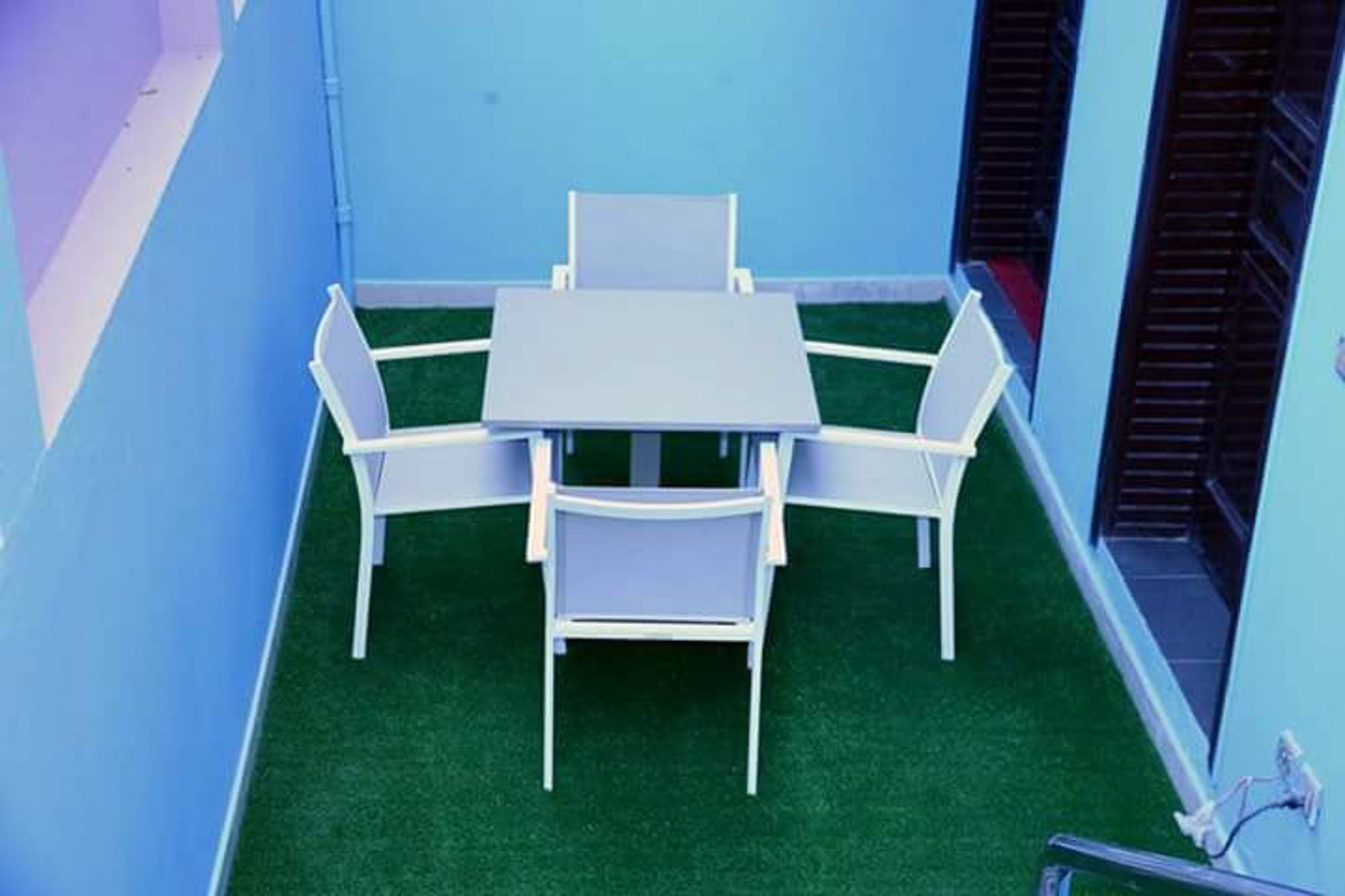 Un pequeño espacio al aire libre con una mesa y sillas, rodeado de paredes azules y césped artificial verde.